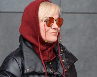 May include: A woman wearing a red knit balaclava with a black and red beaded glasses chain. The balaclava has a long neck and ties at the back. She is wearing a black leather jacket.