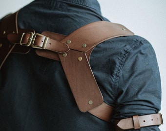 May include: A brown leather camera strap with adjustable buckles and brass accents. The strap is worn over the shoulder and across the chest.
