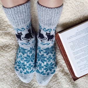 May include: A pair of gray knit socks with a snowflake and cat pattern. The socks have a teal and black design, with a ribbed cuff. The socks are worn with a book open on a white surface.