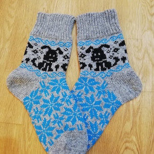 Puede incluir: Un par de calcetines de punto grises con un patrón de copo de nieve azul y negro. Los calcetines tienen un diseño de perro blanco y negro en la parte superior.