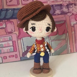 May include: A crocheted doll featuring a brown hat, dark brown hair, and black button eyes. The doll wears a yellow and red checkered shirt with a red neckerchief, a black and white vest, blue pants, and brown boots, standing on a round wooden base.