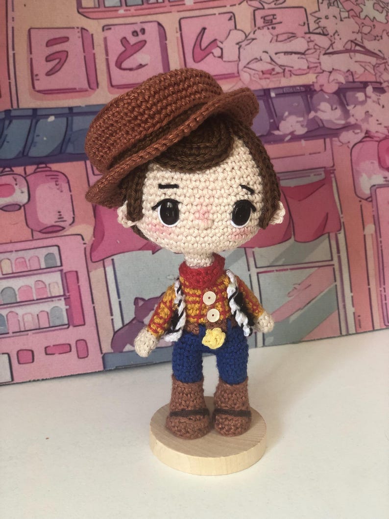May include: A crocheted doll featuring a brown hat, dark brown hair, and black button eyes. The doll wears a yellow and red checkered shirt with a red neckerchief, a black and white vest, blue pants, and brown boots, standing on a round wooden base.