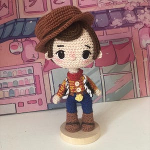 May include: A crocheted doll featuring a brown hat, dark brown hair, and large black eyes. The doll wears a yellow and red plaid shirt with two white buttons, a red neckerchief, blue pants, and brown boots, standing on a circular wooden base.