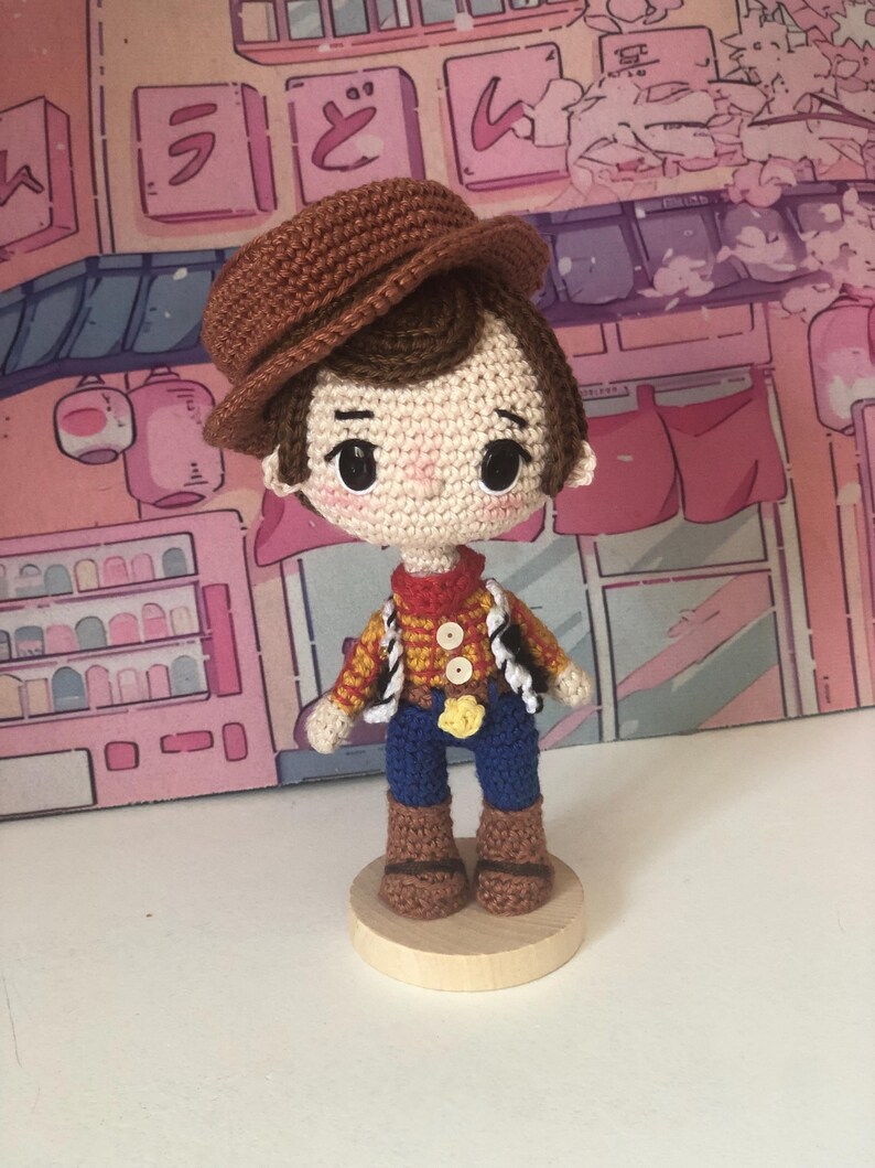 May include: A crocheted doll featuring a brown hat, dark brown hair, and large black eyes. The doll wears a yellow and red plaid shirt with two white buttons, a red neckerchief, blue pants, and brown boots, standing on a circular wooden base.
