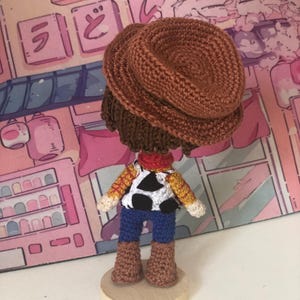 May include: A crocheted doll of a cowboy character wearing a brown hat, a red neckerchief, a yellow and red plaid shirt with a white and black cow-print vest, blue pants, and brown boots, standing on a circular light-colored wooden base.