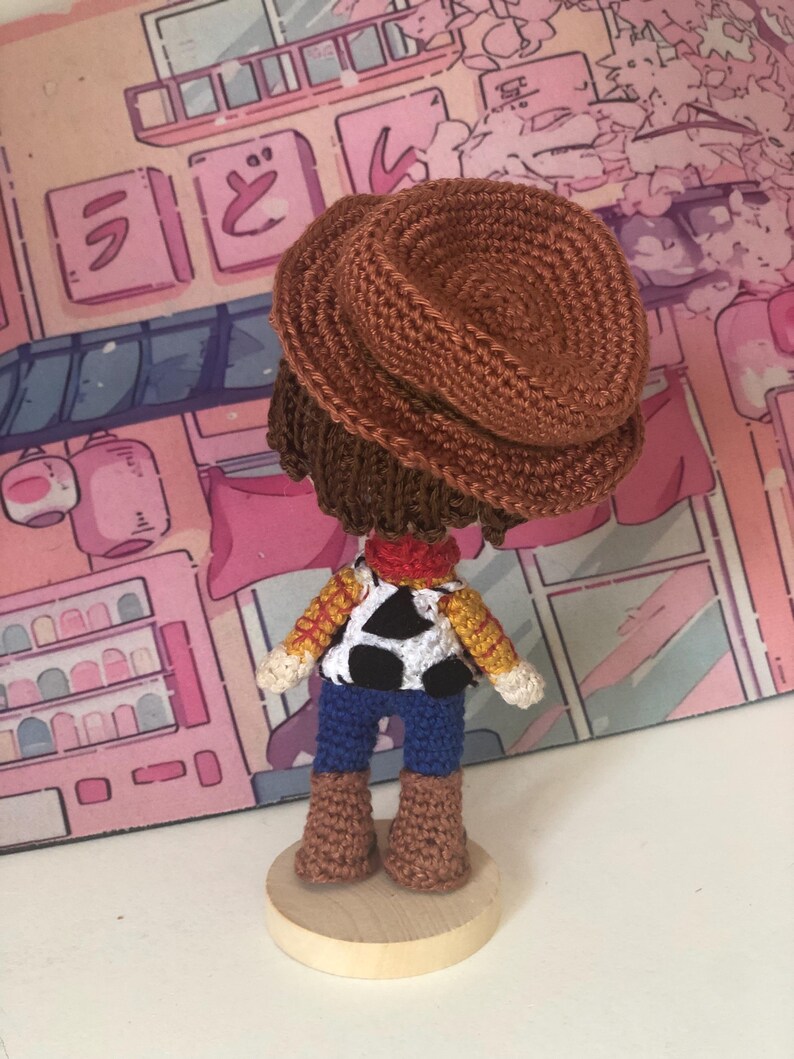 May include: A crocheted doll of a cowboy character wearing a brown hat, a red neckerchief, a yellow and red plaid shirt with a white and black cow-print vest, blue pants, and brown boots, standing on a circular light-colored wooden base.