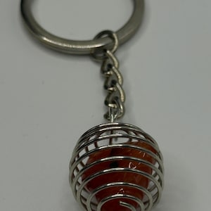May include: A keychain featuring a silver-colored metal cage encasing a round, orange-colored stone. The keychain includes a silver ring and a short chain connecting the ring to the cage.