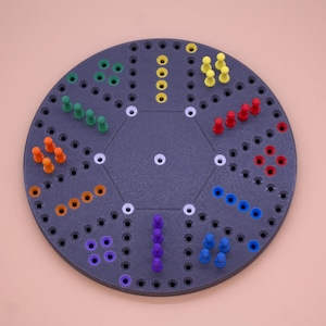May include: A dark gray pegboard game with a circular design. The board features various colored pegs, including green, orange, yellow, red, blue, and purple. The pegs are arranged in patterns around the board, creating a visually engaging game.