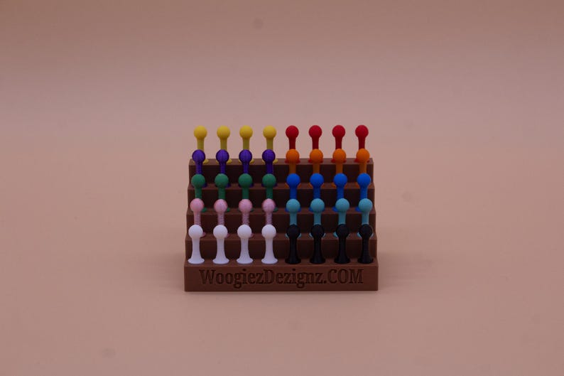 May include: A display stand with a variety of colorful, round-topped, plastic pegs. The stand is made of brown plastic and has the text "WoogiezDezignaz.COM" printed on the front.