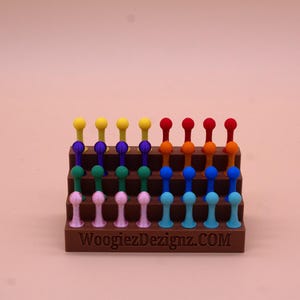 May include: A rainbow-colored set of 24 small plastic pegs arranged in a tiered display stand. The stand is brown and has the text "WoogiezDezignz.COM" printed on the front.