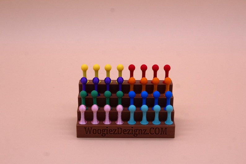 May include: A rainbow-colored set of 24 small plastic pegs arranged in a tiered display stand. The stand is brown and has the text "WoogiezDezignz.COM" printed on the front.