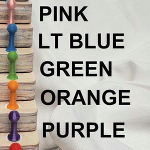 May include: A set of 10 colorful push pins, each with a different color, arranged in a vertical stack on a wooden surface. The colors are black, white, turquoise, pink, light blue, green, orange, purple, red, and yellow. The colors are labeled next to each pin.