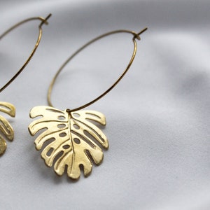 May include: A pair of gold-colored hoop earrings. Each earring features a cutout leaf design, resembling a monstera leaf. The earrings are suspended from a thin, circular hoop. The background is a soft, white fabric.