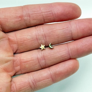 May include: A pair of small gold-coloured stud earrings. One earring is a star shape, and the other is a crescent moon shape. The earrings are displayed on a light background.