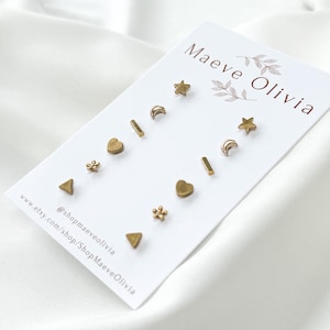 May include: A set of gold-colored stud earrings in various shapes, including stars, moons, hearts, triangles, and floral designs. The earrings are displayed on a white card with the text "Maeve Olivia" and social media information.