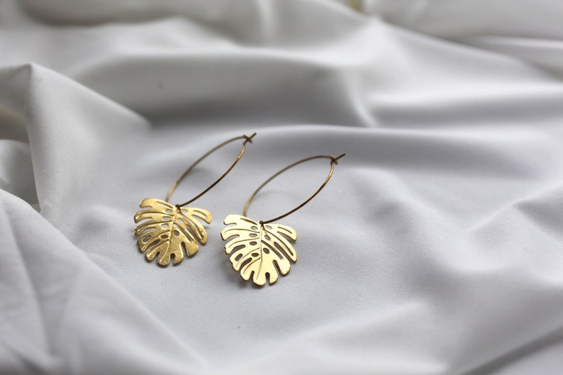 May include: A pair of gold-colored hoop earrings with a monstera leaf design. The earrings are displayed on a white, textured fabric. The leaves have intricate cut-out details, and the hoops are thin and delicate.