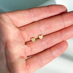 Puede incluir: Un par de pequeños pendientes de botón dorados en forma de corazón. Los pendientes se muestran sobre un fondo claro.