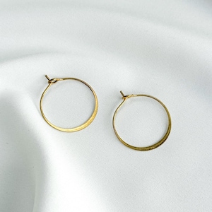 Puede incluir: Dos pendientes de aro dorados. Los pendientes son circulares con una superficie ligeramente texturizada. Los pendientes se muestran sobre un fondo blanco. Una joya sencilla y elegante.