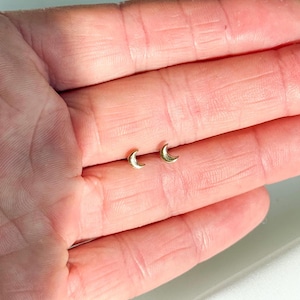 Op de afbeelding: Twee kleine, goudkleurige halve maan oorbellen. De oorbellen worden gepresenteerd tegen een lichte achtergrond. De oorbellen zijn klein en delicaat, met een eenvoudig, elegant ontwerp. De oorbellen zijn waarschijnlijk gemaakt van goud of een goudkleurig metaal.