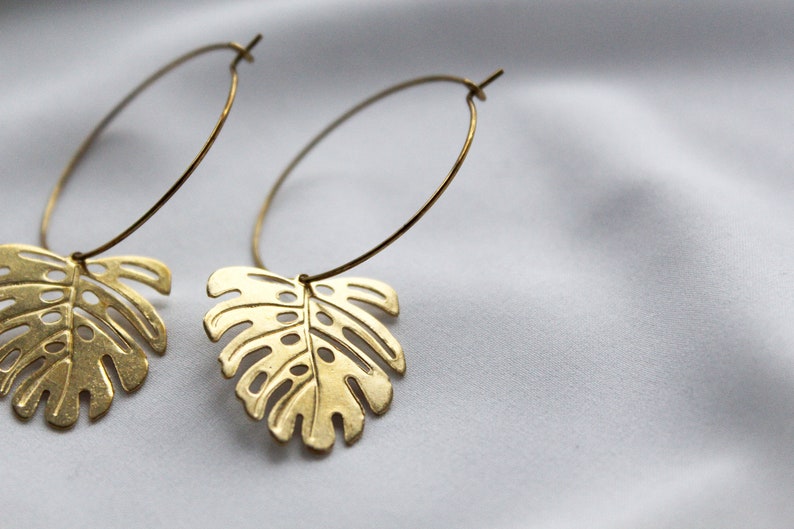 May include: A pair of gold-colored hoop earrings. Each earring features a cutout leaf design, resembling a monstera leaf. The earrings are suspended from a thin, circular hoop. The background is a soft, white fabric.