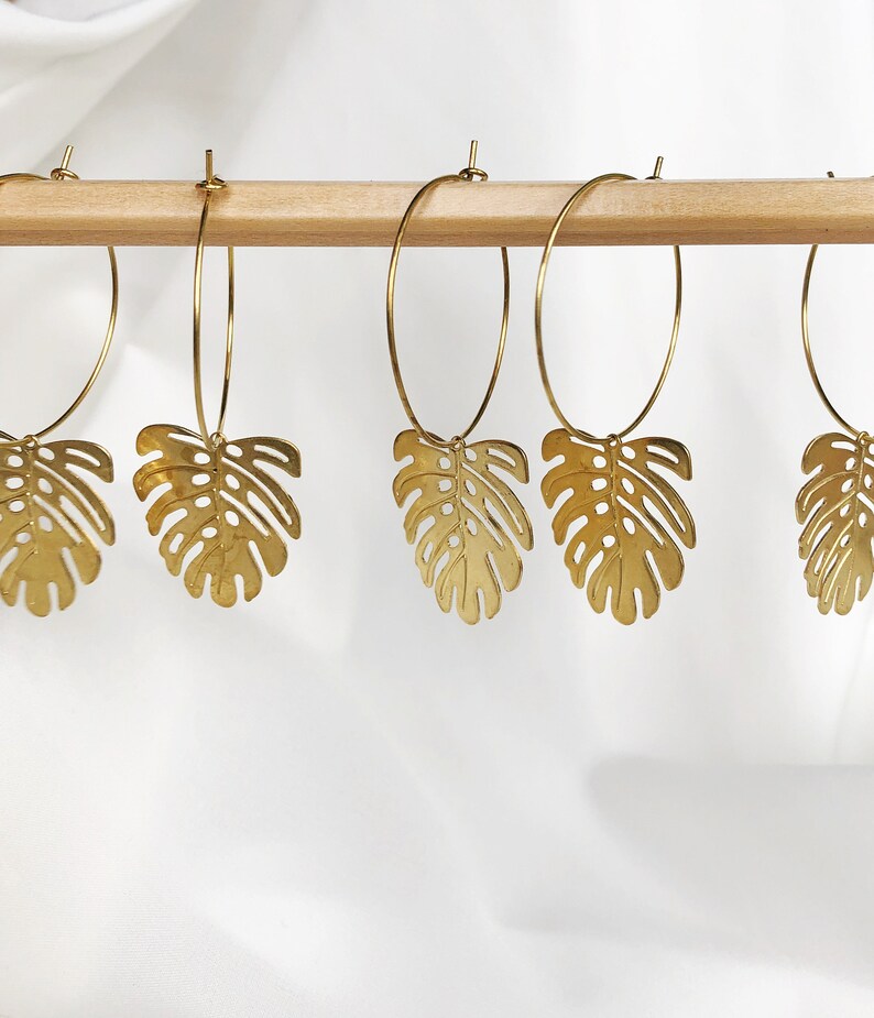 May include: Gold-colored hoop earrings with a monstera leaf charm. The earrings are hanging on a wooden rod against a white background. The earrings have a thin hoop design and a detailed leaf pendant.
