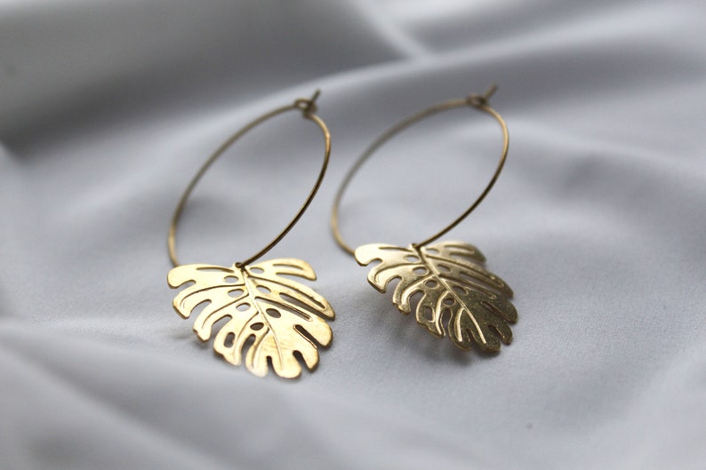 May include: A pair of gold-colored hoop earrings. Each earring features a cutout monstera leaf charm suspended from a thin, circular hoop. The earrings are displayed against a soft, white background.