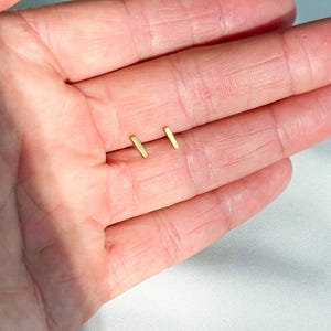 May include: A pair of small, gold-coloured bar stud earrings. The earrings have a simple design, each featuring a straight, rectangular bar shape. The earrings are displayed on a light background.