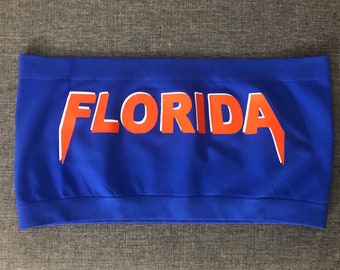 May include: A blue strapless top with the word "FLORIDA" printed in orange with a white outline in a metal band font.