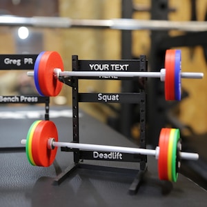 Peut inclure: Un rack de musculation en métal noir avec trois supports pour haltères. Chaque support est équipé d'une barre avec des plaques de poids colorées. Les supports sont étiquetés "Bench Press", "Squat" et "Deadlift".