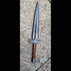 May include: A silver and brown sword with a double-edged blade. The hilt features a decorative design and a brown leather-wrapped grip. The sword is a prop or decorative item.