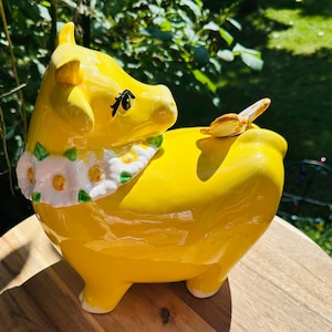 May include: A bright yellow ceramic cow figurine featuring a glossy finish, a white daisy flower garland around its neck, and a small butterfly perched on its back. The cow has painted black eyelashes and a smooth, rounded body shape.