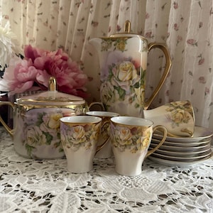 Puede incluir: Un juego de té vintage con un diseño floral. El juego incluye una tetera, un azucarero, una jarra de crema y cuatro tazas y platillos. El diseño floral presenta rosas blancas y hojas verdes sobre un fondo morado claro. El juego está adornado con detalles dorados.