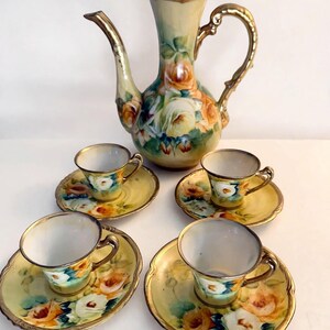 May include: A porcelain tea set consisting of a tall teapot with a lid and four matching cups and saucers. Each piece features a hand-painted floral design with yellow and white roses, green foliage, and gold trim on the rims and handles.