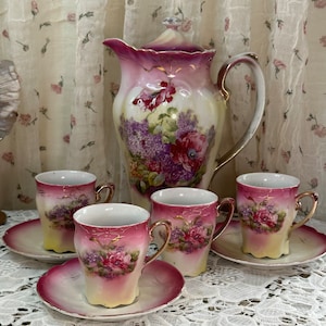 Könnte beinhalten: Ein Vintage-Tee-Set mit Blumenmuster. Das Set enthält eine Teekanne, vier Tassen und vier Untertassen. Die Teekanne hat eine rosa-weiße Basis mit einem Blumendesign in Violett-, Rosa- und Grüntönen. Die Tassen und Untertassen haben ein ähnliches Design mit einer rosa-weißen Basis und einem Blumendesign in Violett-, Rosa- und Grüntönen.