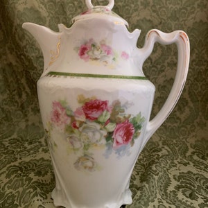 May include: A white porcelain teapot with a floral design featuring pink and white roses. The teapot has a gold trim and a green band around the middle. The teapot has a lid with a heart-shaped handle.