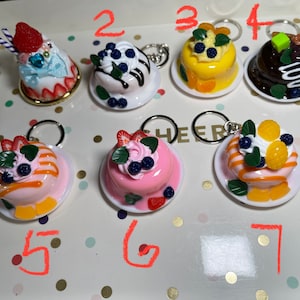 May include: Ten miniature cake keychains with different toppings, including strawberries, blueberries, and orange slices. The cakes are on white plates with silver key rings.