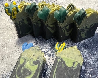 May include: A set of five green and black swirl soap bars with yellow and blue decorative accents. The soap bars are arranged in a row on a gray background.
