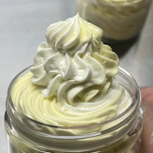 May include: A glass jar filled with a thick, creamy, yellow body butter. The butter is topped with a swirl of white whipped cream.
