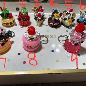 May include: Nine colorful Christmas-themed cake charms with keychains. The charms are decorated with frosting, sprinkles, and candy. The charms are numbered 1 through 9.