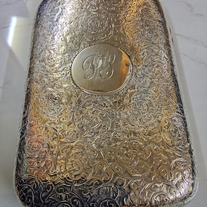 May include: A rectangular silver-toned metal case featuring an intricate, embossed scrollwork pattern across its surface. A smooth, circular central medallion is engraved with the stylized, interconnected cursive letters "PS" in the middle.