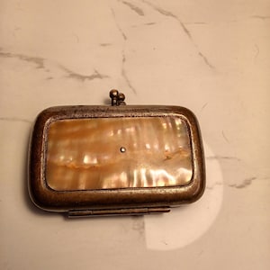 May include: An antique rectangular purse with a mother-of-pearl front and a tarnished metal frame. The purse has a small clasp and a hinged bottom. The mother-of-pearl has a warm, iridescent glow.