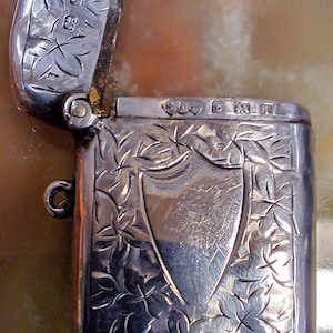 May include: A silver vesta case with a hinged lid, featuring an intricate floral engraving surrounding a central blank shield-shaped cartouche. The metal surface displays detailed leaf patterns and visible hallmarks along the top edge of the body.
