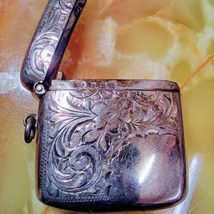 May include: A silver vesta case with an open hinged lid, featuring intricate floral and scrollwork engravings on the front surface. The metal container includes a small side loop and visible hallmarks stamped along the top edge of the body.