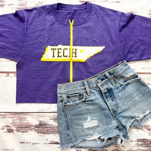 May include: Purple cropped t-shirt with a yellow zipper and a white graphic of the state of Tennessee with the word "TECH" on it. The t-shirt is paired with light blue denim shorts.