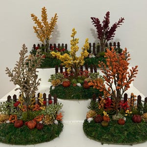 May include: Five miniature garden displays featuring artificial trees in shades of yellow, orange, red, and brown. Each display includes a small brown picket fence, green mossy ground cover, and tiny orange pumpkins arranged at the base.