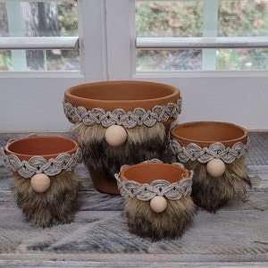 Op de afbeelding: Vier terracotta potten met een dwergontwerp. Elke pot heeft een bruin viltbaard, een houten neus en een beige touwversiering rond de bovenkant.