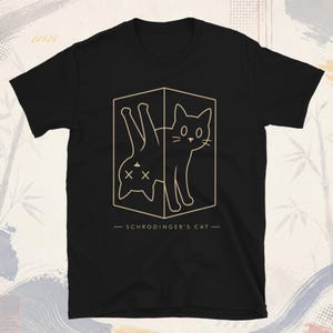 以下が含まれることがあります： 箱に入った猫のゴールドのラインアートが描かれた黒の半袖Tシャツ。猫は2つの状態で表現されており、片方は目を開けて立っており、もう片方は逆さまになって目がX印になっています。テキストには「SCHRODINGER'S CAT」と書かれています。