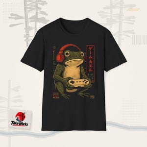 May include: A black t-shirt featuring a graphic of a green frog wearing red headphones and holding a game controller. The design includes Japanese text, the words Player 1, and geometric accents in red and beige, creating a detailed illustration.