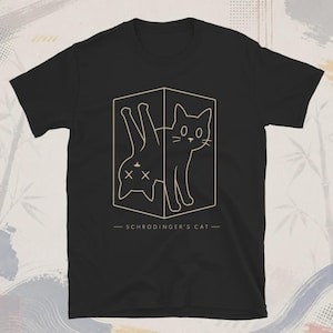 Könnte beinhalten: Ein schwarzes Kurzarm-T-Shirt mit einer goldenen Linienzeichnung einer Katze in einer Box. Die Katze ist in zwei Zuständen dargestellt, einmal aufrecht und einmal auf dem Kopf stehend mit X-förmigen Augen. Der Text SCHRODINGER'S CAT steht unter der Box.