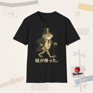 Könnte beinhalten: Ein schwarzes Kurzarm-T-Shirt mit einer Grafik eines Frosches, der einen Zylinder, einen Gehrock und eine Weste trägt und einen Gehstock hält. Unter dem Frosch steht der japanische Text 蛙が帰った。 Das Design ist in gedeckten Beige- und Dunkeltönen.